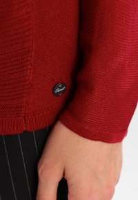 Pull en maille rouge avec une finition texturée et des poignets côtelés. Présente une étiquette circulaire noire avec le nom de la marque sur l'ourlet.