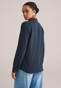 Tmavomodrá košeľa s dlhými rukávmi a bielymi tenkými prúžkami, so zvýrazneným golierom, voľného strihu s gombíkovými manžetami. Nosí sa s svetlomodrými širokými džínsami.