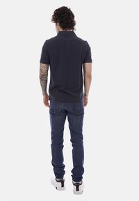 Homme aux cheveux bouclés et au bras droit tatoué, portant un polo noir, un jean bleu foncé et des baskets blanches, vu de dos.