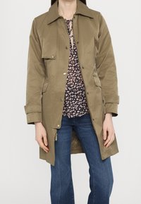 Trench-coat kaki avec col, fermeture à pressions à l'avant et poches latérales ; porté sur un chemisier fleuri et un jean bleu. Texture lisse.