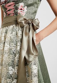 Robe verte à motifs floraux avec ceinture en satin olive. Le surjupe en dentelle présente une broderie complexe blanche et dorée.