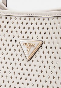 Sac à main beige avec des perforations en forme de diamant et un badge logo métallique triangulaire Guess USA en or et blanc au centre.