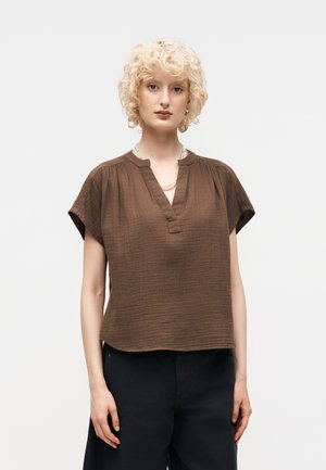 Άτομο με ξανθά, σγουρά μαλλιά που φοράει χαλαρό καφέ κοντομάνικο μπλουζάκι και μαύρο παντελόνι, στέκεται μπροστά σε απλό ανοιχτό φόντο.