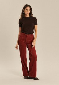 Pantalons évasés en velours côtelé rouge foncé pour femmes, avec une taille haute, des côtes verticales et une fermeture à bouton. Associés à un haut ajusté de couleur sombre.