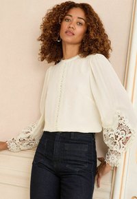 White blouse with intricate lace details on puffed sleeves, paired with dark denim high-waisted jeans. Smooth fabric with delicate accents.