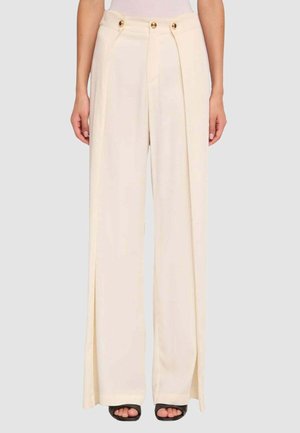 Donna che indossa pantaloni crema a vita alta e gamba larga con bottoni dorati e spacchi laterali, abbinati a tacchi neri aperti.