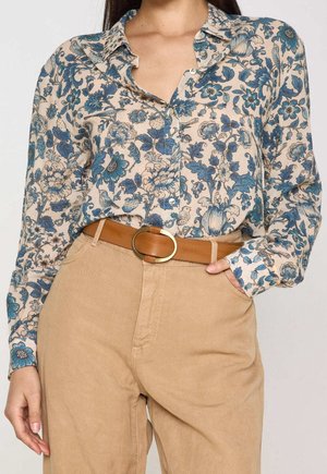 Femme portant une chemise beige à fleurs bleues boutonnée, rentrée dans un pantalon beige avec une ceinture marron à boucle dorée arrondie.