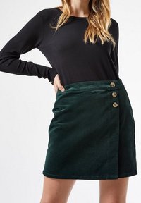 Grön kordverkskjol med en omslagdesign och tre metallknappar som accent vid midjan. Mjuk textur och rak fåll.