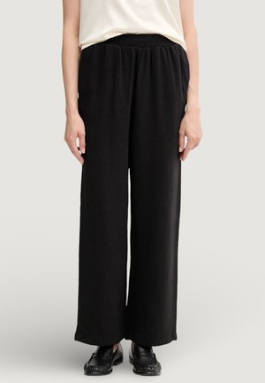 Pantaloni neri a gamba larga realizzati in tessuto di trama. Presentano una cintura elastica e arrivano fino alle caviglie. Abbinati a mocassini neri.