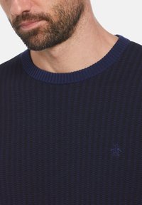 Pull en tricot bleu marine avec une texture côtelée, doté d'un col rond et d'un petit logo brodé sur la poitrine.