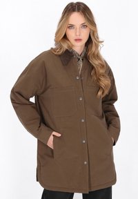Kvinde med langt bølget blond hår iført en brun knapjakke med store lommer og en skjorte med paisley-mønster under.
