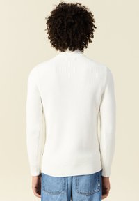 Maglione bianco a costine con colletto alto, maniche lunghe aderenti e orlo dritto. Indossato con pantaloni di jeans azzurri chiari.