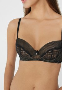 Czarny koronkowy biustonosz z przejrzystymi paskowanymi miseczkami, delikatnym kwiatowym wzorem oraz małą kokardką z ozdobnym wisiorkiem na środku. Regulowane ramiączka.