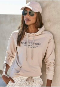 Sudadera beige con el texto "NATURE VIBES", cordones con acentos metálicos, combinada con pantalones claros a rayas de cebra y una gorra a juego.