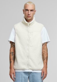 Gilet en polaire blanc cassé avec col montant, fermeture éclair intégrale et poche avant. Tissu texturé avec un aspect doux, porté sur un t-shirt blanc.