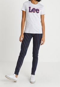 Camiseta gris con logo rojo de "Lee", combinada con jeans de rayas verticales en azul oscuro. El modelo lleva zapatillas blancas. Un atuendo simple y casual.