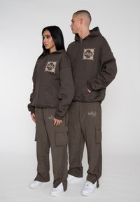 Brune zip-up hættetrøjer med dekorative lapper og matchende cargo bukser, der har lommer og broderede detaljer, suppleret med hvide sneakers.