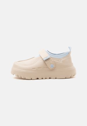 Beige slip-on sko med tyk gummisål, justerbar stofrem med knap og lyseblåt inderfoer på hvid baggrund.