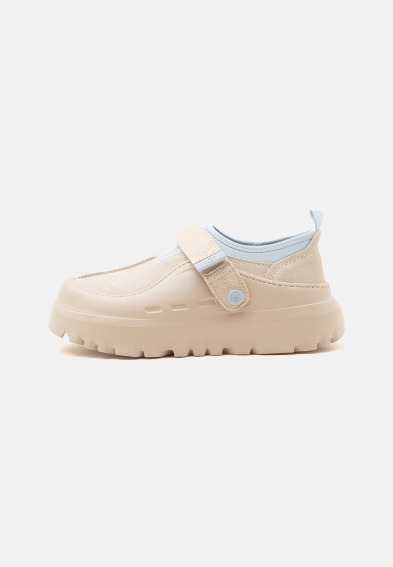 Chaussure beige à enfiler avec semelle épaisse en caoutchouc, bride en tissu ajustable avec bouton et doublure intérieure bleu clair sur fond blanc.
