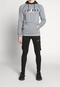 Sudadera gris con detalles en negro, que presenta un gran logotipo. Pantalones cargo negros con bolsillos y bordados en dorado. Zapatillas negras completan el conjunto.
