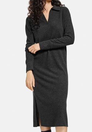 Kvinde iført en langærmet, mørkegrå, knælange strikkjole med krave og sideslids, stående mod en ensfarvet lys baggrund.