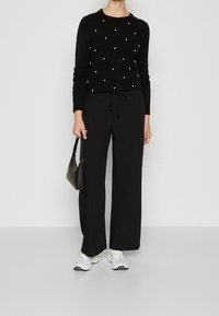 Svart stickad tröja med pärldetaljer, matchad med svarta vida byxor och vita sneakers. Modellen håller en liten svart handväska.