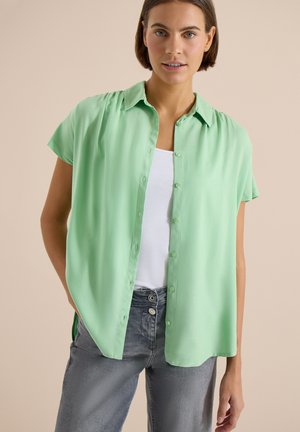 Kvinde iført en lysegrøn kortærmet skjorte med knapper over en hvid top og grå jeans mod en beige baggrund.