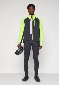 Conjunto de ciclismo que incluye un cortavientos negro y verde neón con paneles de malla, combinado con pantalones ajustados negros y un casco negro.