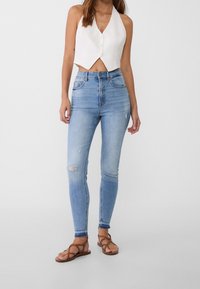 Blaue Denim-Skinny-Jeans mit distressed Knie-Details, kombiniert mit einem weißen, kurz geschnittenen Haltertop. Getragen mit braunen, flachen Sandalen mit Riemen.