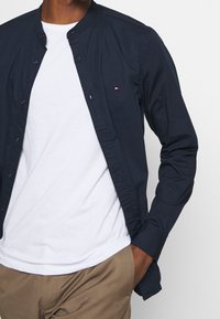 Camicia button-up blu navy con colletto, decorata con un piccolo logo rosso, bianco e blu. Indossata sopra una maglietta bianca e abbinata a pantaloni beige.