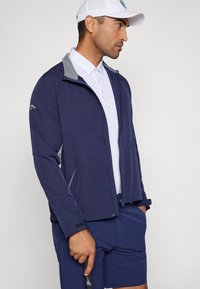 Marinejakke med grå detaljer, hel glidelås, to frontlommer, lett stoff, kombinert med en hvit polo og marineblå shorts.