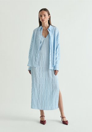 Mujer con vestido sin mangas texturizado en azul claro combinado con camisa abierta de manga larga a juego y tacones puntiagudos burdeos, de pie sobre un fondo liso.