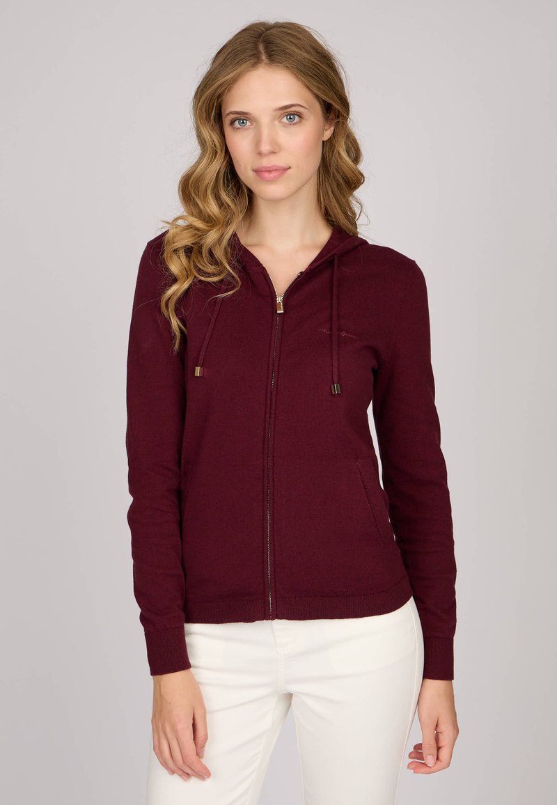 Sweat à capuche zippé bordeaux avec un cordon de serrage, des poignets et un ourlet côtelés, et des poches avant. Texture lisse et coupe ajustée.