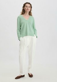 Mintgrüner V-Ausschnitt-Pullover aus lockerem Strick, kombiniert mit weißen, gefalteten Hosen und braunen Wildleder-Ballerinas. Weiche Textur und entspannte Passform.