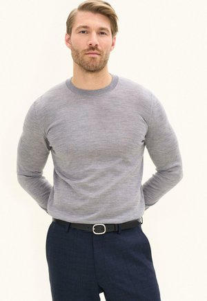 Mann trägt einen hellgrauen Langarmpullover, dunkelblaue Hose und schwarzen Gürtel, steht mit den Händen hinter dem Rücken vor einfarbigem Hintergrund.