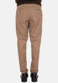 Uomo che indossa pantaloni eleganti beige e scarpe eleganti nere, in piedi con le gambe ravvicinate su uno sfondo semplice.