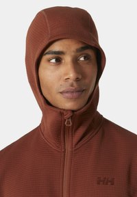 Chaqueta marrón de textura con cremallera y capucha ajustada, además de un alto collar. Cuenta con un logotipo sutil en el pecho y está fabricada con un tejido estructurado.