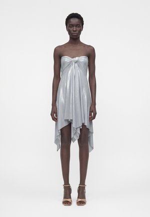 Femme debout portant une robe métallique argentée sans bretelles avec un nœud sur le devant et un ourlet asymétrique, associée à des sandales à talons hauts beiges.