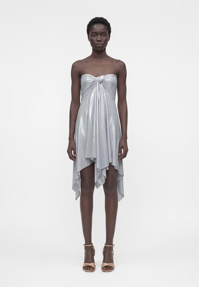 Mulher em pé vestindo um vestido metálico prateado sem alças com nó frontal e bainha assimétrica, combinado com sandálias de salto alto bejes.