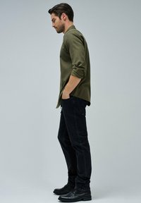 Camicia verde oliva con bottoni, maniche arrotolate, abbinata a jeans neri aderenti e stivali in pelle nera, mostrata di profilo.