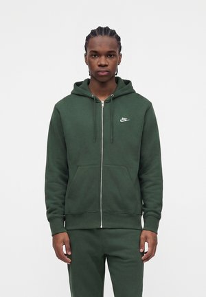 En ung mand står vendt fremad iført en mørkegrøn Nike hættetrøje med lynlås og matchende bukser mod en ensfarvet hvid baggrund.