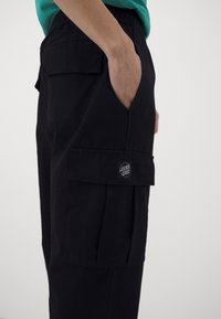 Mustat cargo-housut, joissa on joustava vyötärönauha, sivutaskut ja lahkeessa oleva merkkilogo. Valmistettu kestävästä puuvillamateriaalista.