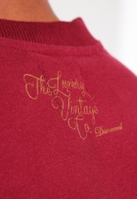Marooninvärinen puuvillainen collegepaita, jossa on resorimainen kaulus ja kullanvärisellä langalla kirjailtu "The Luxury Vintage Co." -teksti niskan takana.