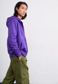 Hanorac violet dintr-un material moale, cu buzunar tip cangur, asortat cu pantaloni cargo verzi, prevăzuți cu buzunare laterale și o croială lejeră.