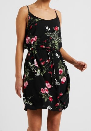 Kobieta ubrana w czarną, bezrękawnikową sukienkę z cienkimi ramiączkami i czerwono-różowo-białym kwiatowym wzorem, przewiązaną w talii paskiem z tkaniny.