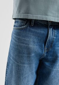 Niebieskie jeansy z denimu o klasycznym kroju, wyposażone w zamek błyskawiczny, pięć kieszeni i efekt sprania. Połączone z jasnoszarą koszulą.