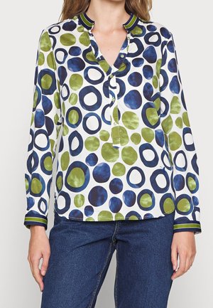 Frau trägt eine langärmelige Bluse mit grünen und blauen kreisförmigen Mustern und gestreiften Kragen und Manschetten, kombiniert mit blauen Jeans.