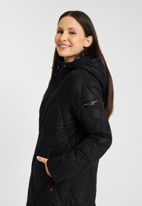 Zwarte gewatteerde pufferjack met een capuchon, zijvakken en een diagonaal patroon. Heeft een merketiket op de mouw en een soepele textuur.