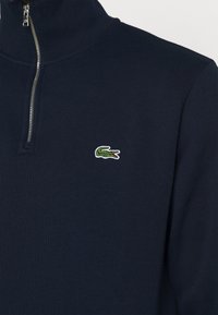 Tamsiai mėlynas zip-neck megztinis, pagamintas iš tekstūruoto mezgimo audinio, su mažuoju žaliu ir raudonu įsiūtu krokodilo logotipu ant krūtinės.
