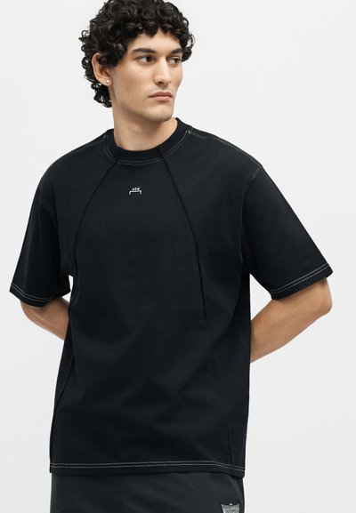 Άνδρας με σγουρά μαλλιά που φοράει μαύρο κοντομάνικο μπλουζάκι με λευκές ραφές και ένα μικρό λευκό λογότυπο στο στήθος.